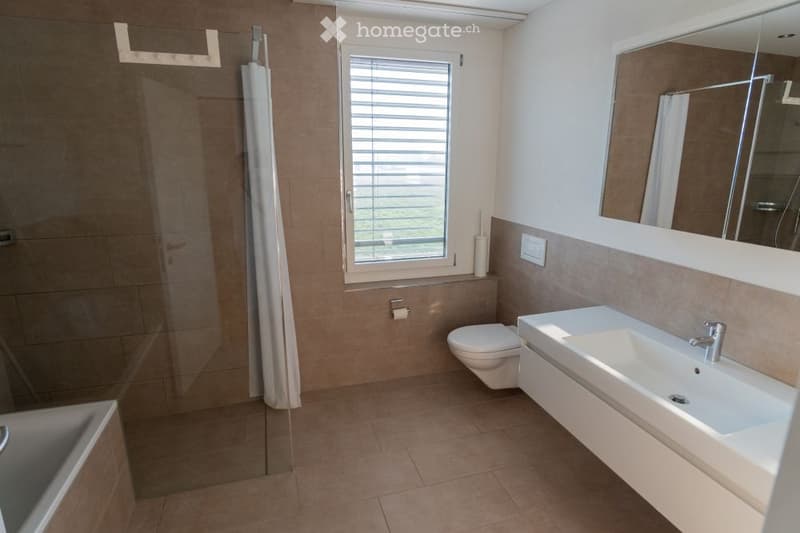 Modern bathroom with a large mirror, a white sink, a toilet, a bathtub, and a shower curtain. The room features beige tiles and a window with blinds.