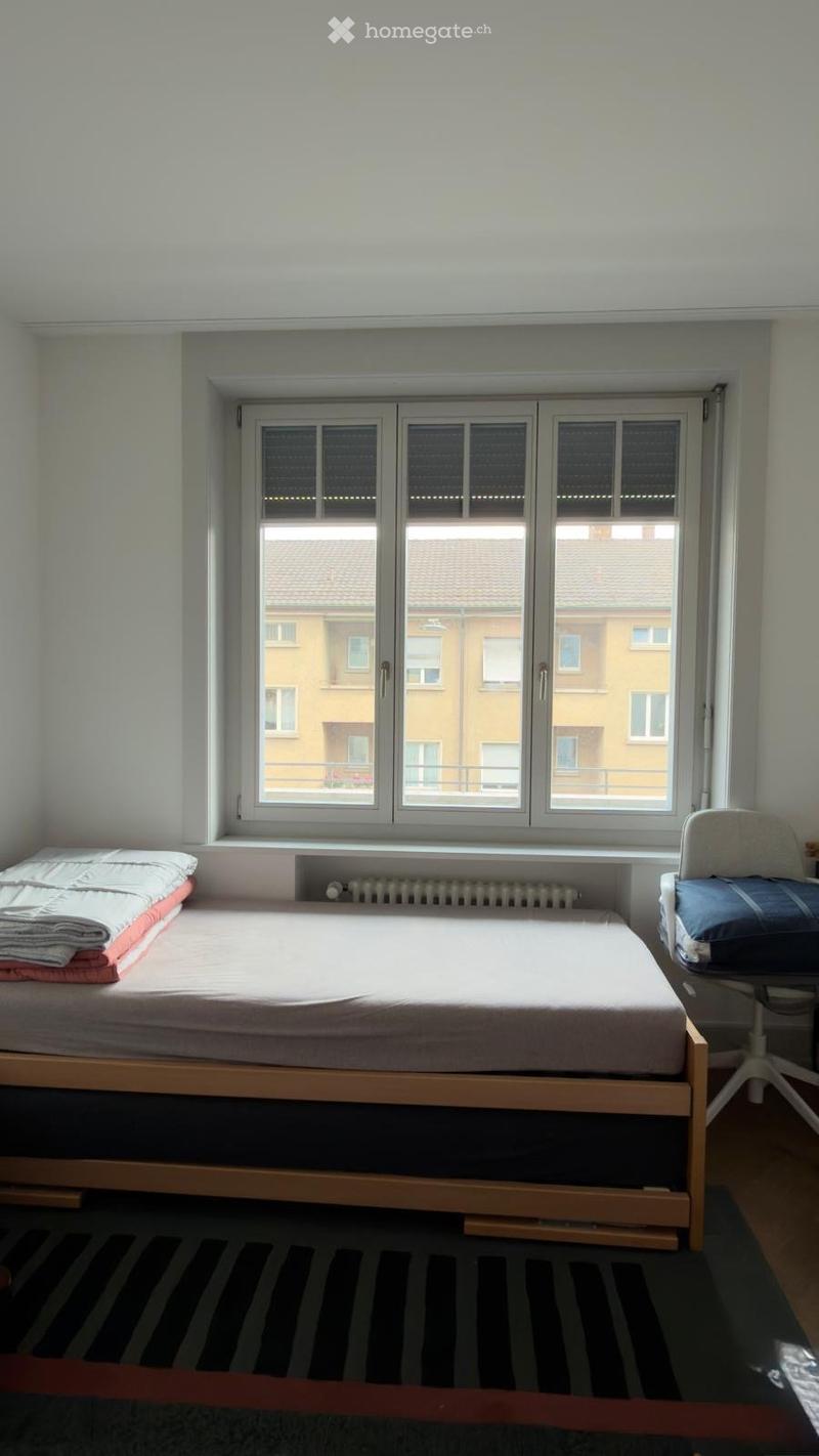 Minimalist bedroom with a single bed, a small chair, and a radiator beneath a large window overlooking a residential area.
