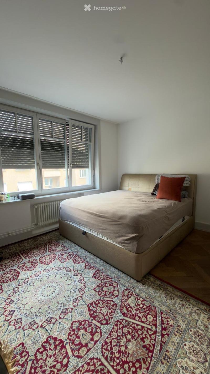 A well-lit bedroom with a large bed, patterned rug, and large windows with blinds.