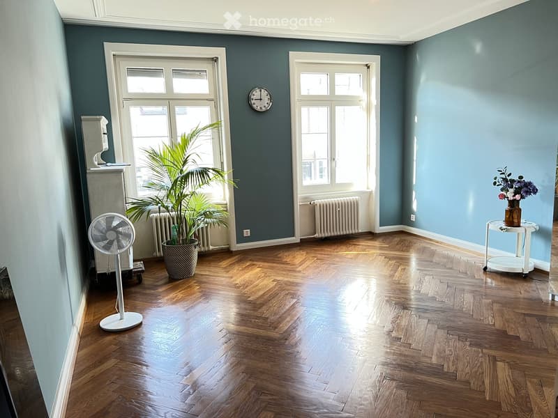 Empty room with wooden flooring, blue walls, large windows, a fan, a potted plant, and a small table with a vase of flowers.