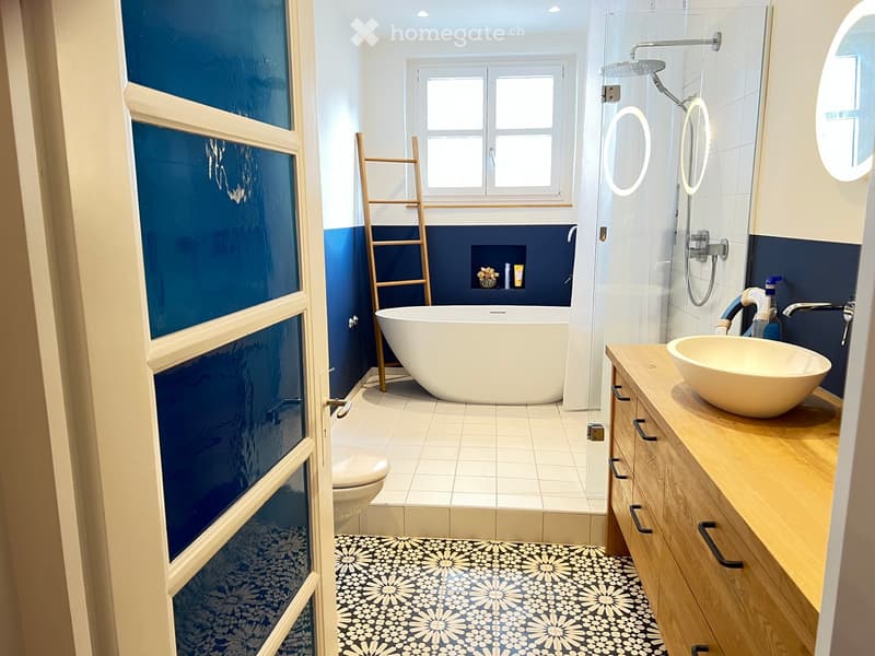 Modern bathroom with a freestanding bathtub, a sink with a wooden vanity, a toilet, a glass-enclosed shower, and a wooden ladder leaning against the wall. The walls are painted in a two-tone design with dark blue lower half and white upper half. The floor