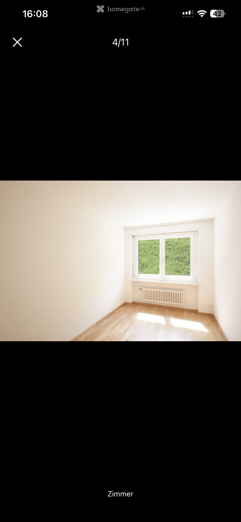 Empty room with light wooden flooring, white walls, and a large window with white frame showing greenery outside. A radiator is visible below the window.
