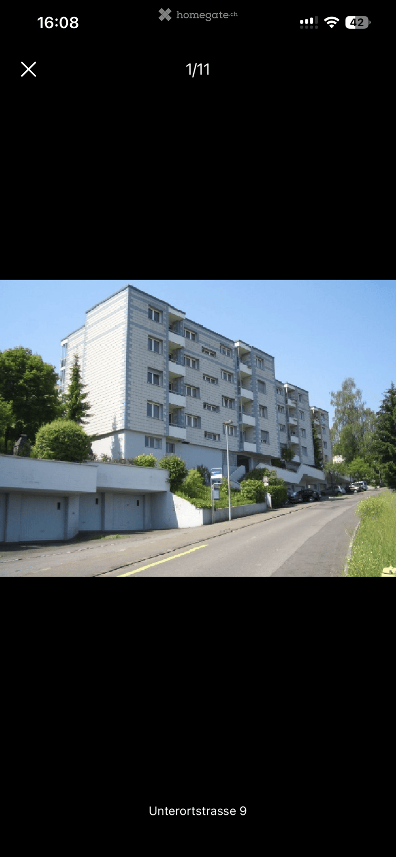 Multi-story residential building with white facade, multiple windows, and balconies. A lower-level garage is visible, with a paved road in front and greenery on the sides.