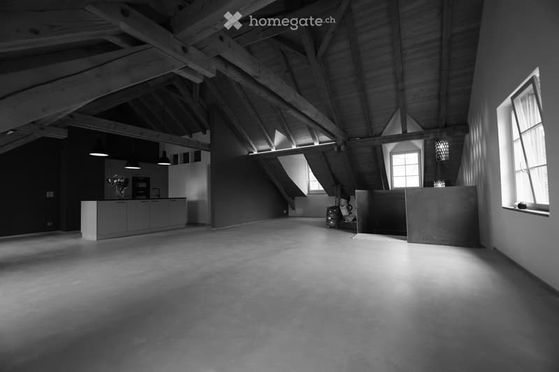An empty, spacious room with exposed wooden beams and a high, slanted ceiling. The room features large windows allowing natural light to enter, and a small kitchen area is visible in the background.