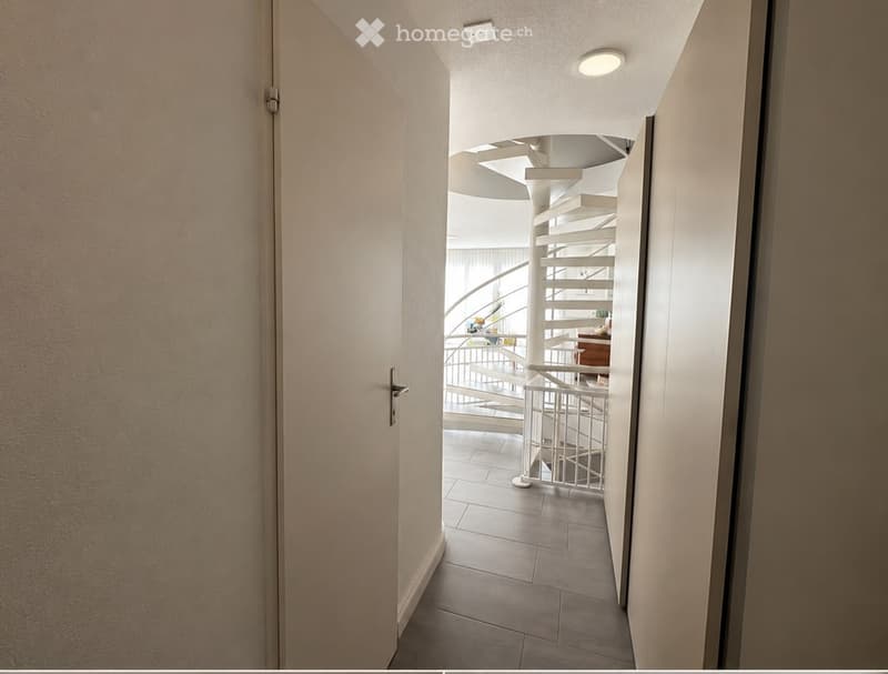 A modern hallway with a curved staircase visible in the background, featuring white walls, tiled flooring, and recessed lighting.