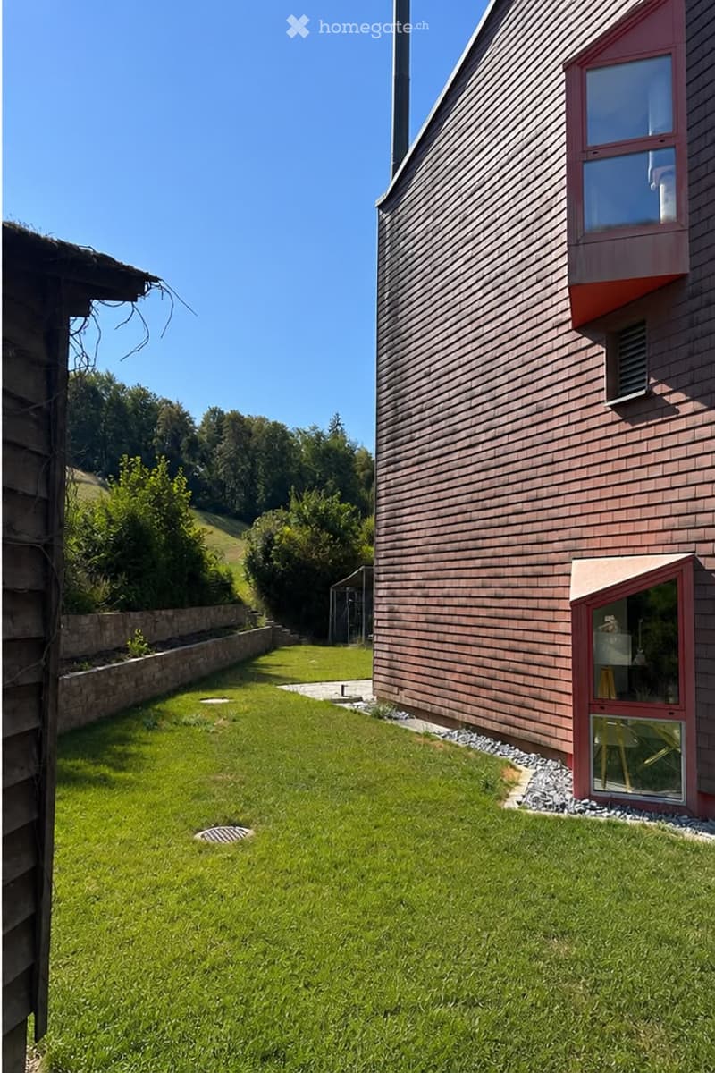 Well-maintained lawn with a stone border, a wooden house with a red-framed window and door, and a clear blue sky in the background.