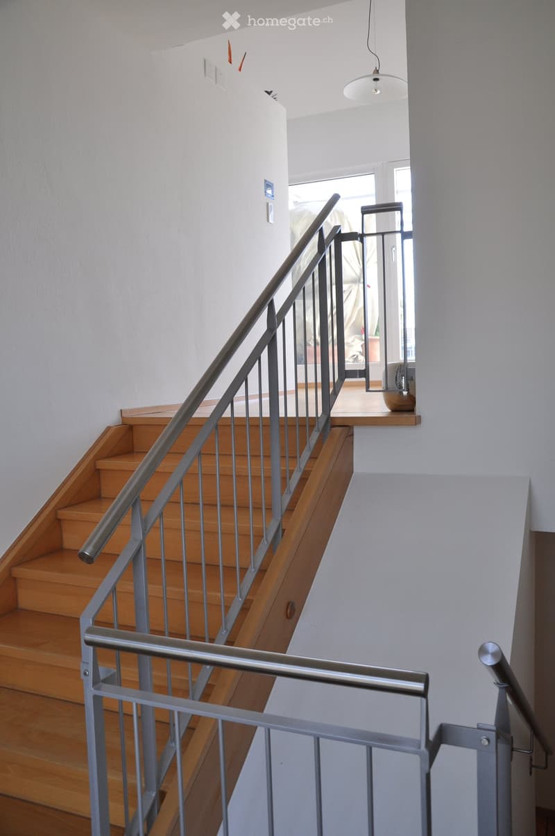 Wooden staircase with metal railing, white walls, and a hanging light fixture. The landing area shows a glimpse of a room with a potted plant and a piece of furniture.
