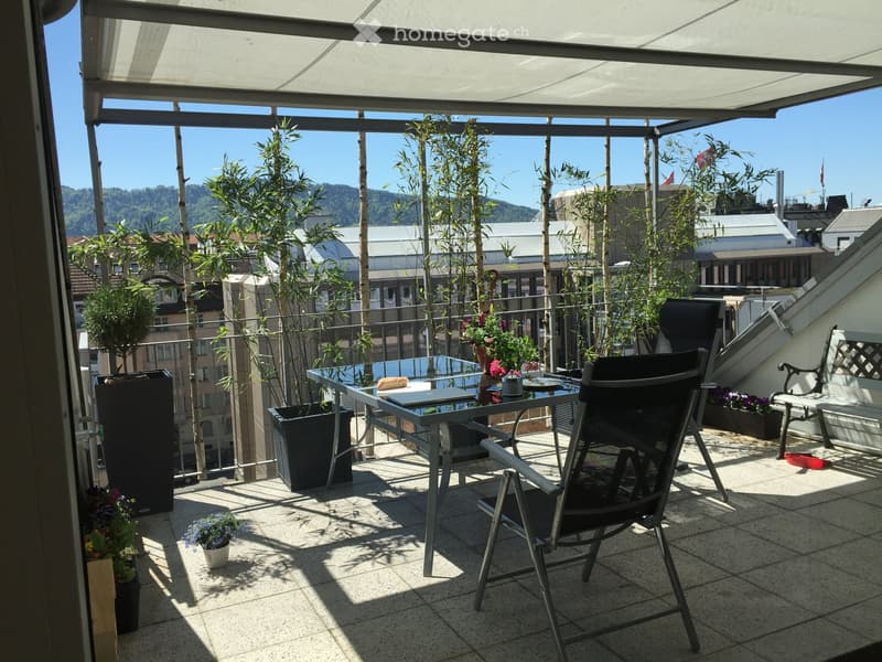 Balcony with a glass table, folding chair, bench, and several potted plants, overlooking a cityscape with trees and buildings.