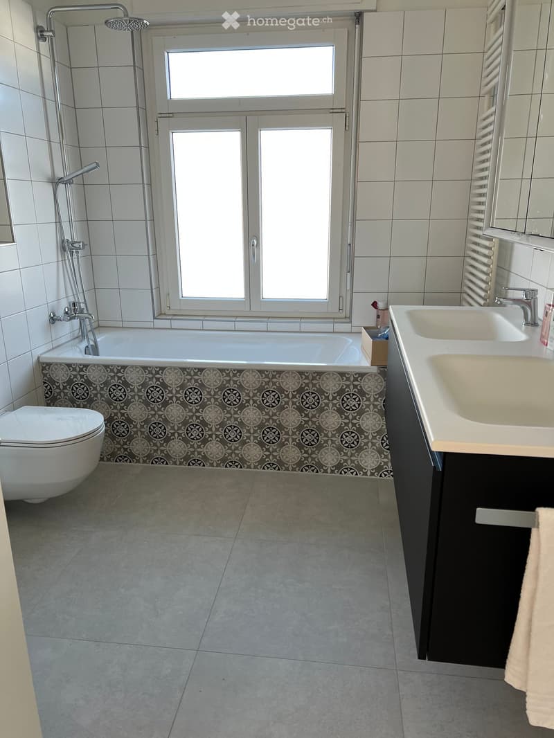 Modern bathroom with a bathtub, toilet, double sink, and a large window. The bathtub features a decorative tile border, and the sink has a modern faucet. A towel is visible on the cabinet.