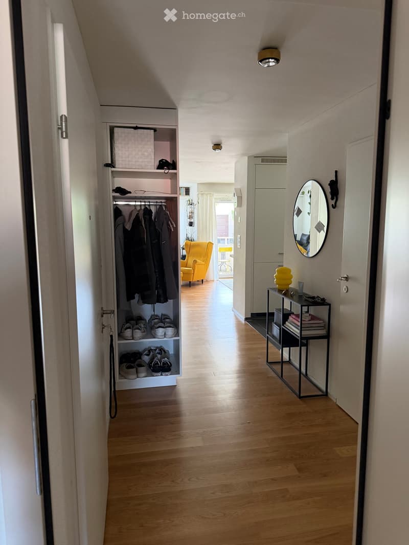 A well-lit hallway with wooden flooring, built-in shoe and coat storage, a small black side table with books and a decorative item, and a round mirror on the wall.