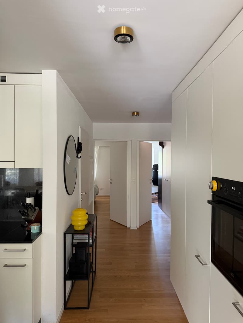Modern hallway with light wooden flooring, white walls, and built-in white cabinets. Features a small black side table with a yellow vase, a mirror on the wall, and an oven on the right side.
