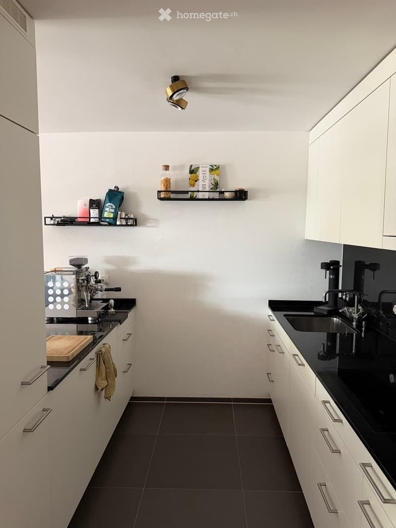 Modern kitchen with white cabinets, black countertops, and stainless steel fixtures. Features a coffee machine, sink, and shelves with various items.