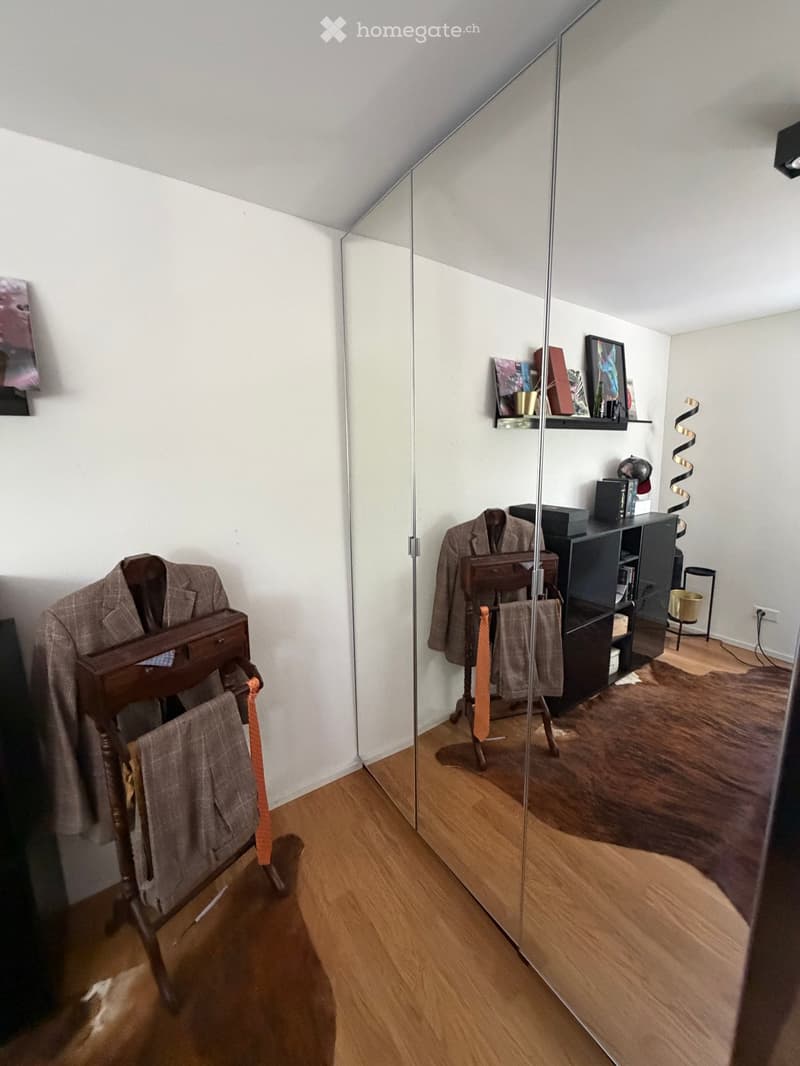 A well-lit room with wooden flooring, a large mirror, a wardrobe, a shelf with books and decorative items, and a small table with a chair.