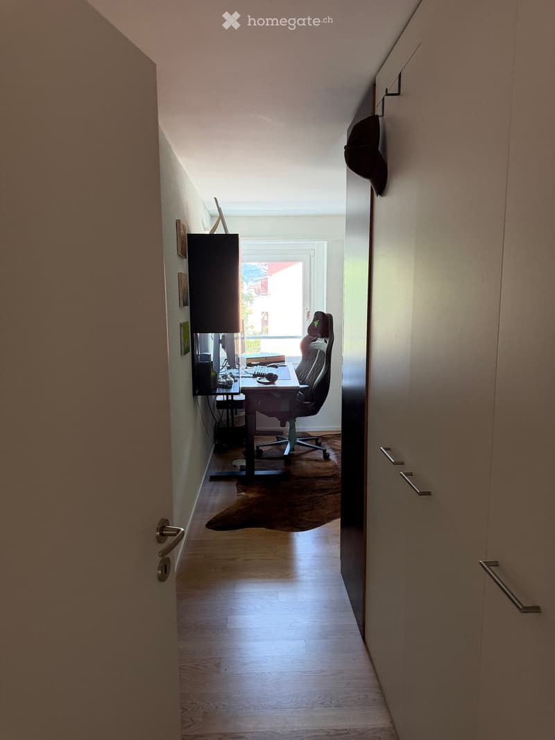 A narrow, well-lit room with wooden flooring, a desk setup including a computer, keyboard, mouse, and a chair, with a lamp on the desk and a window letting in natural light.