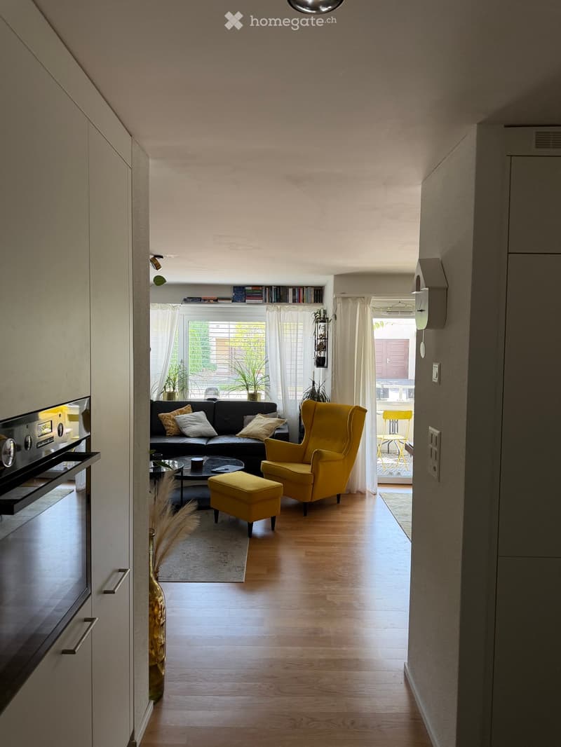 Modern living room with hardwood floors, large windows, black leather sofa, yellow armchair, ottoman, coffee table, and decorative vase.
