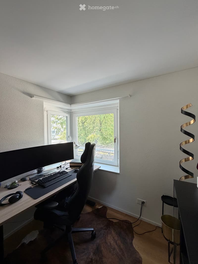 Modern, well-lit room with a large window, a desk with a laptop, keyboard, mouse, and screen, a comfortable chair, and a small table lamp. The room features a sleek, contemporary design with a wooden floor and a stylish, curved wall decoration.
