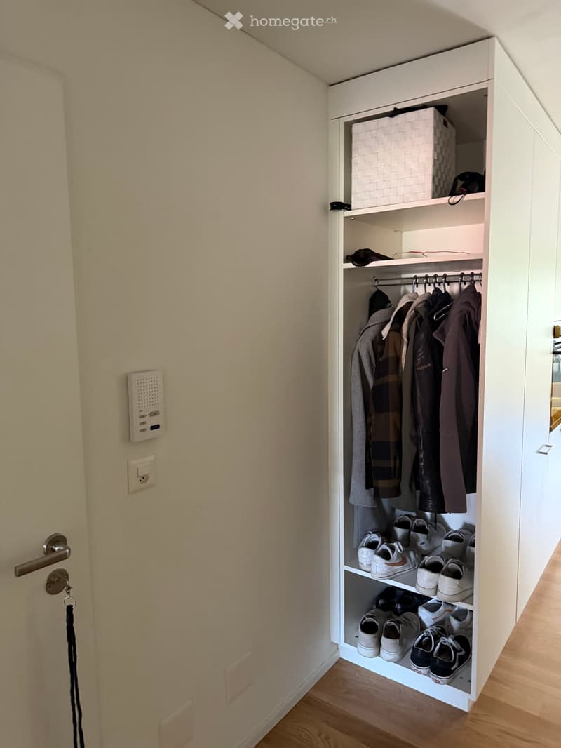 A built-in closet with multiple shelves and hanging space, containing shoes and clothing. The closet is white and has a modern design, with a door featuring a silver handle and a keyhole.