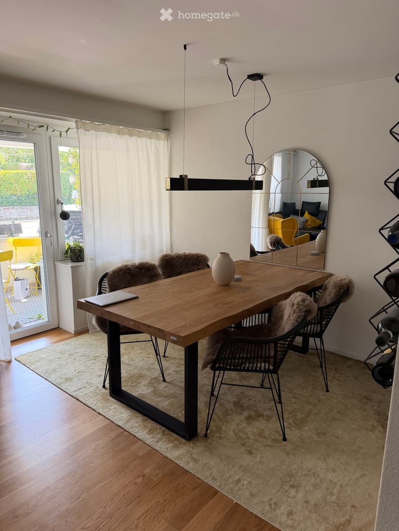Modern dining area with a wooden table, black metal chairs with fur cushions, a laptop, a vase, and a large mirror. The space features a pendant light fixture and a view of a garden through large windows.