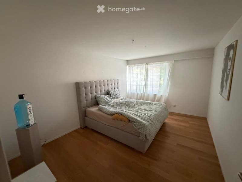 A modern bedroom with a tufted headboard bed, pillows, a blanket, a blue bottle on a nightstand, and a framed picture on the wall. The room features wooden flooring and large windows with white curtains.