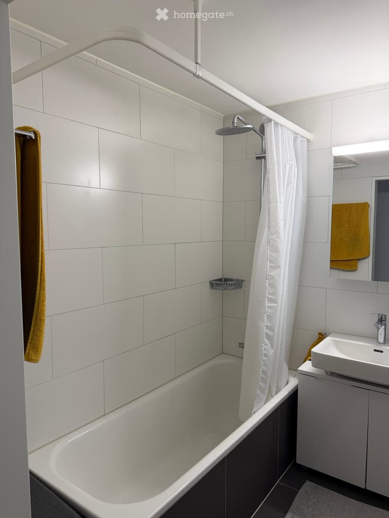 Modern bathroom with a white bathtub, shower, sink, and mirror. White tiles and a white shower curtain are visible.