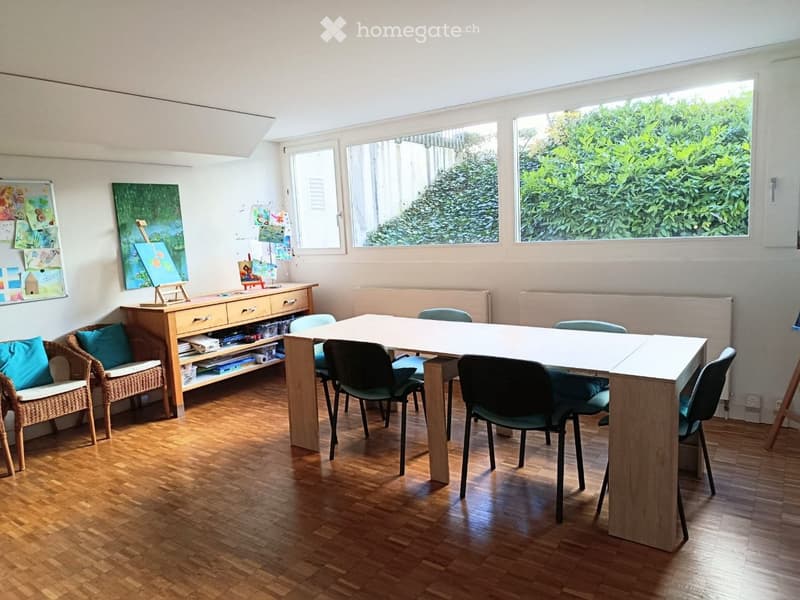 Bright room with wooden flooring, large windows, a long table surrounded by chairs, a wicker sofa with cushions, a wooden cabinet, and several artworks displayed on the walls.