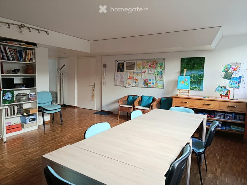 Bright room with wooden flooring, large white table surrounded by light blue chairs, wicker chairs with blue cushions, bookshelf with books and decorative items, easel with painting, and artwork displayed on walls.