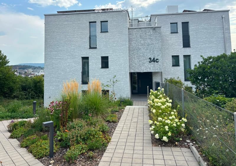 Modern two-story building with grey facade, large windows, and a balcony on the upper level. The entrance is flanked by a small garden area with various plants and flowers, and a paved walkway leads to the door.