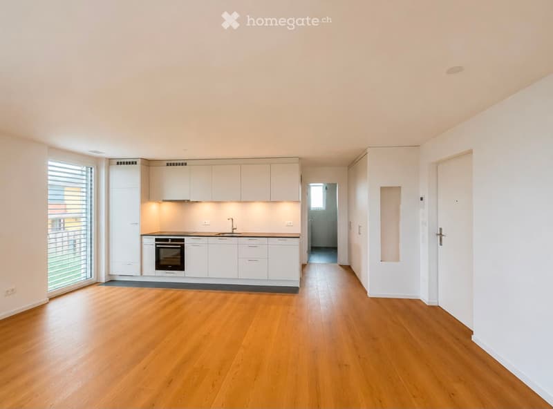 Bright, unfurnished room with light wooden flooring, white walls, and a white kitchen area featuring cabinets, a sink, and an oven. The room has large windows with blinds and a door leading to another space.