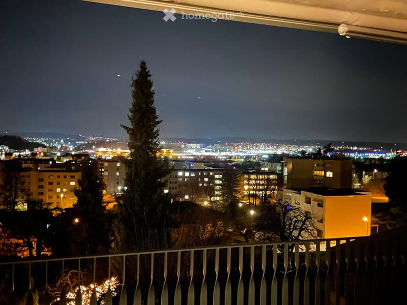 Nighttime view from a balcony showing a cityscape with illuminated buildings, trees, and a distant airport.