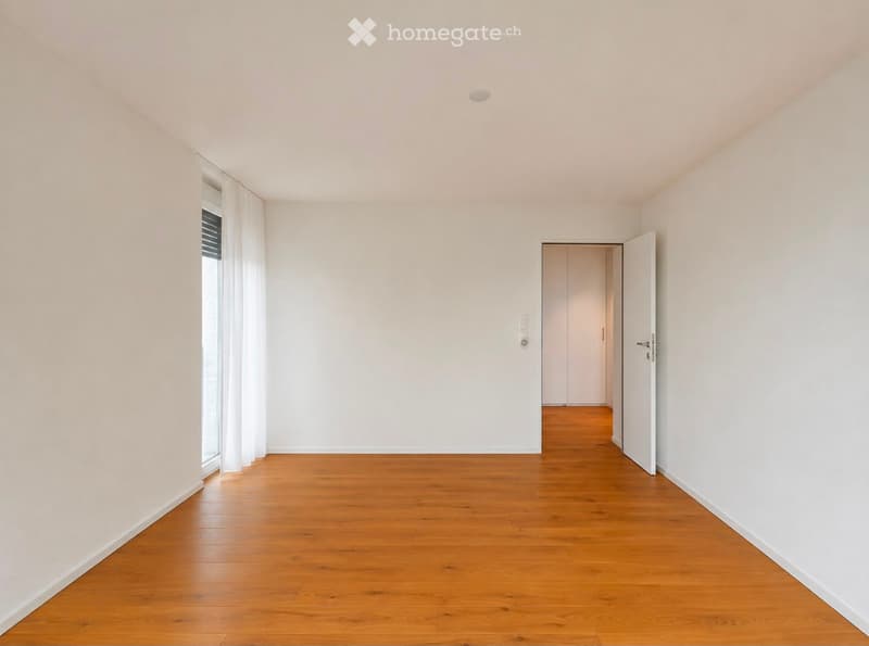 Empty room with light-colored walls, wooden flooring, and a large window with white curtains. An open door leads to another room.