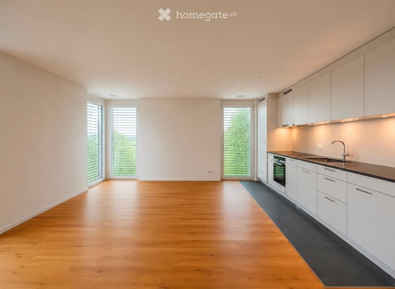 Empty kitchen with white cabinets, wooden floor, and large windows with blinds. No appliances or furniture are present.
