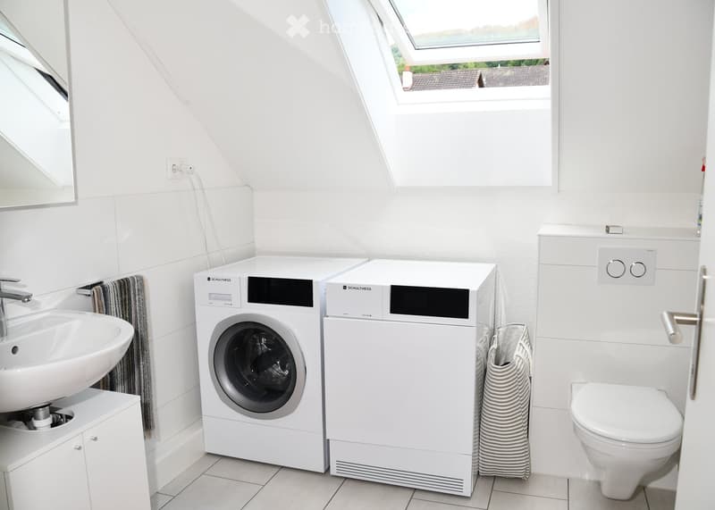 Modern bathroom with white tiles, a sink, a mirror, a washing machine, a dishwasher, and a toilet. The room features a skylight window and a small cabinet under the sink.