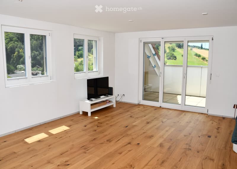 Bright living room with wooden flooring, large windows, and a sliding glass door leading to a balcony. A TV is placed on a white stand.