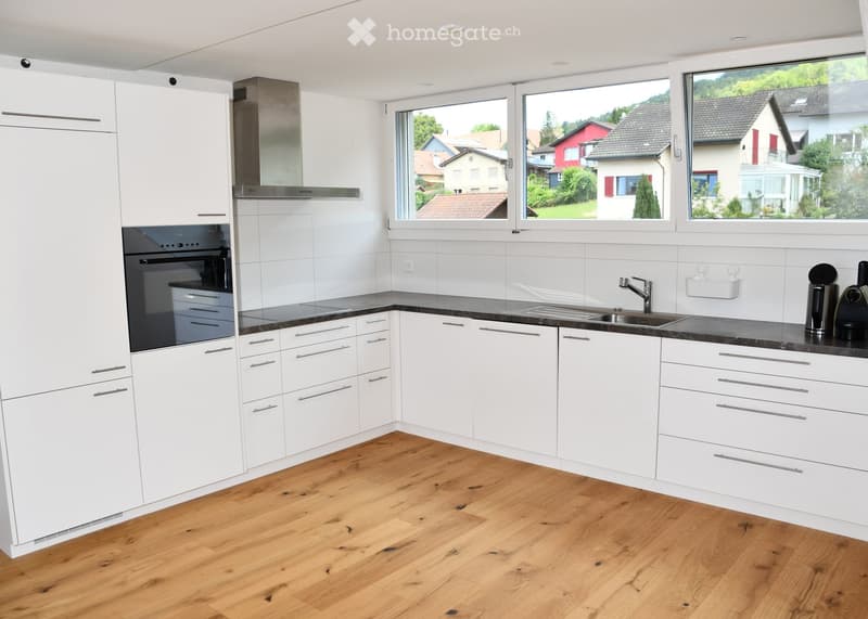 Modern kitchen with white cabinets, wooden flooring, and large windows overlooking a residential area.