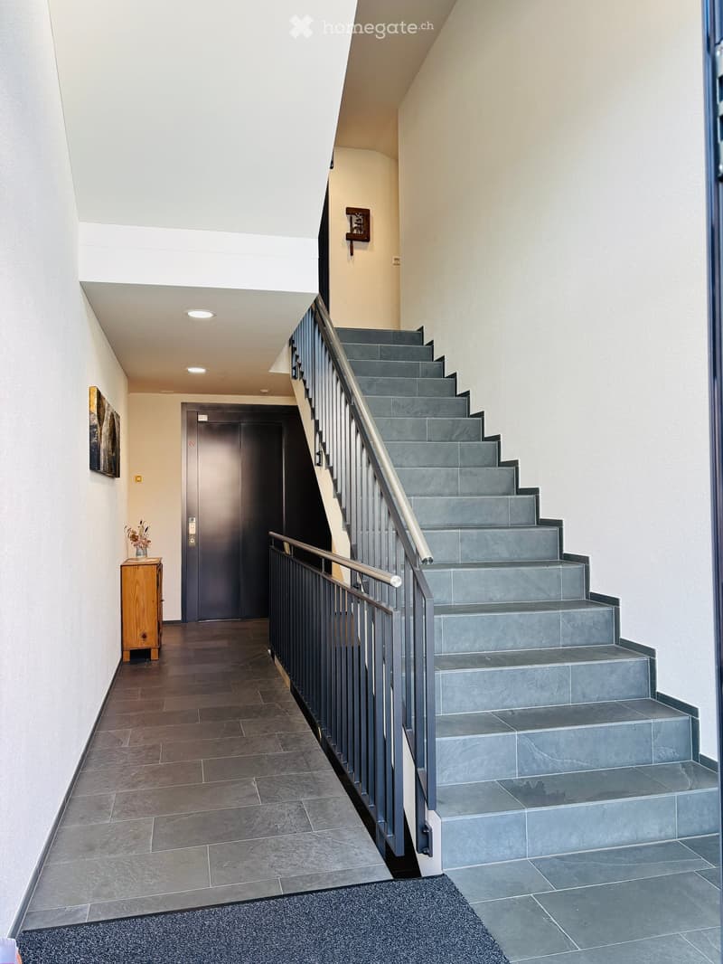 Modern hallway with a staircase leading upwards, featuring grey tiled steps, a metal railing, and a wooden side table with a vase of flowers. The walls are painted in neutral tones, and the floor is covered with dark tiles.