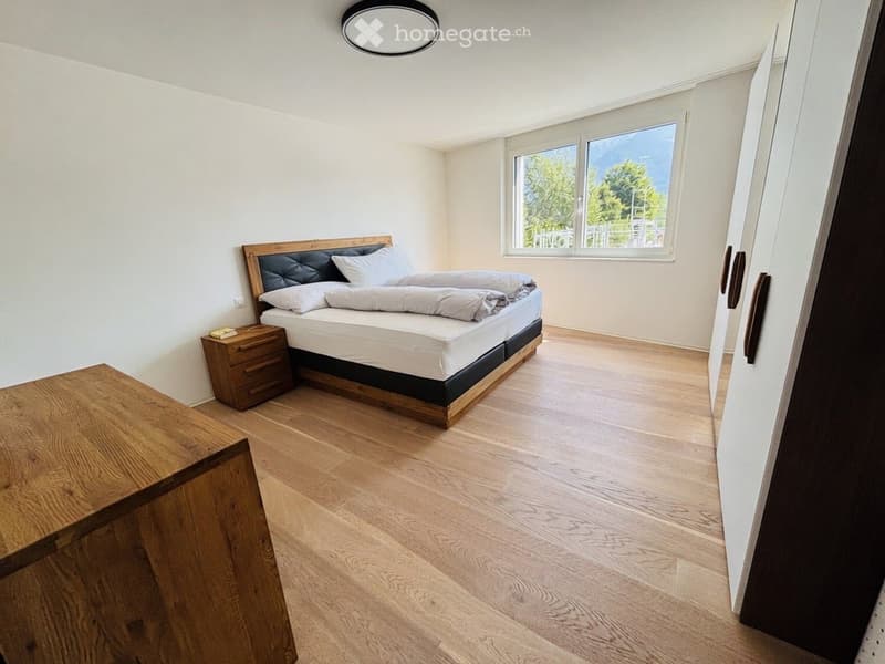 Bright bedroom with wooden flooring, large window offering a view, a bed with white bedding, a nightstand, and a wooden table.