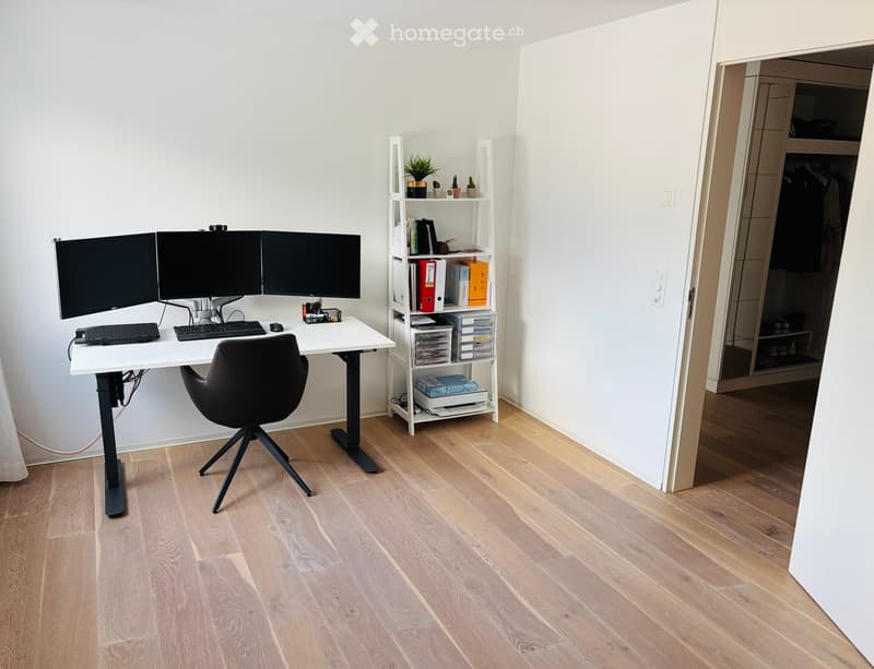 A modern, well-lit home office with a standing desk, ergonomic chair, dual monitors, keyboard, mouse, and a white shelving unit holding books, files, and small plants. The room features light wooden flooring and white walls.