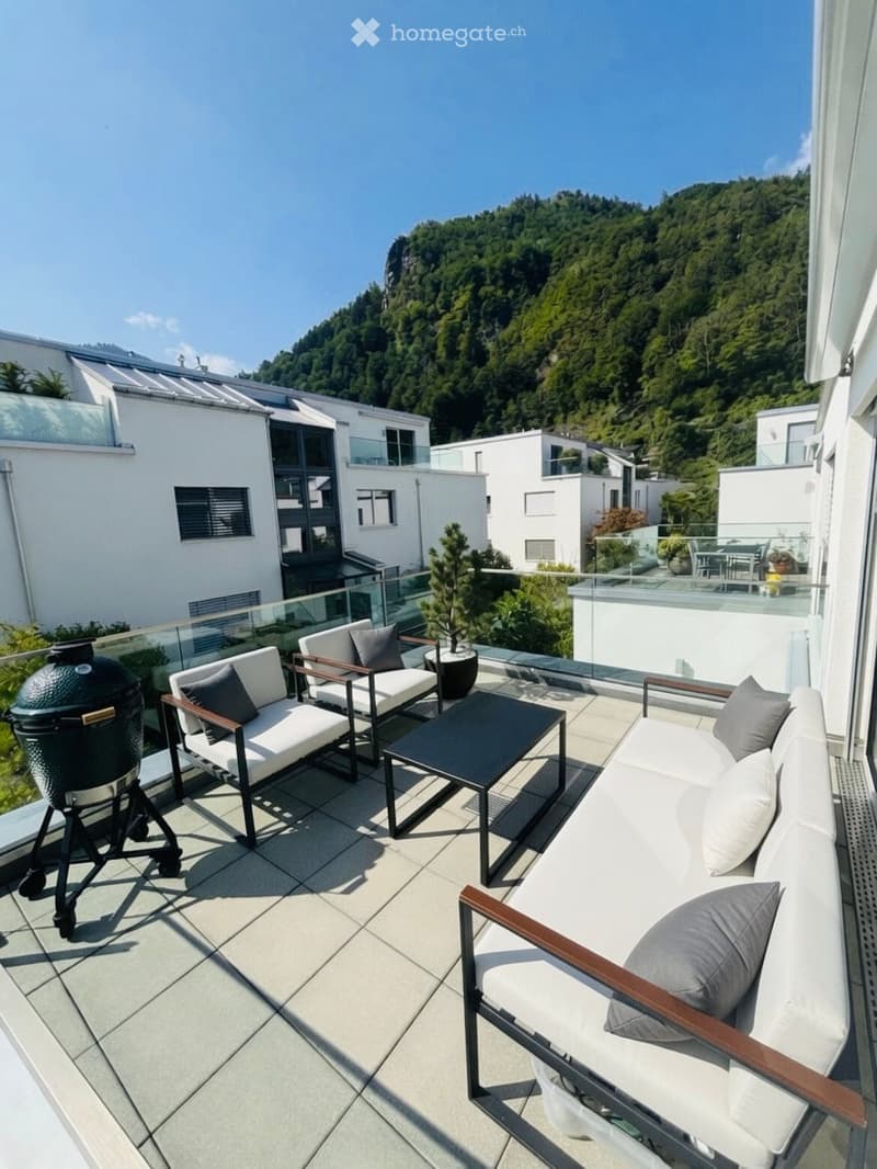 Modern rooftop terrace with white furniture, a grill, and a scenic mountain view.