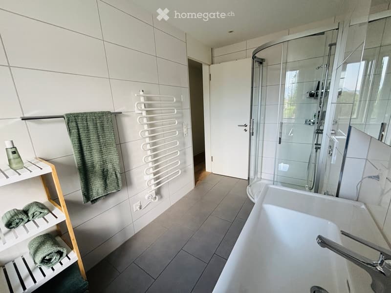 Modern bathroom with a bathtub, glass-enclosed shower, white towel rack, and wooden shelves holding towels and a bottle.