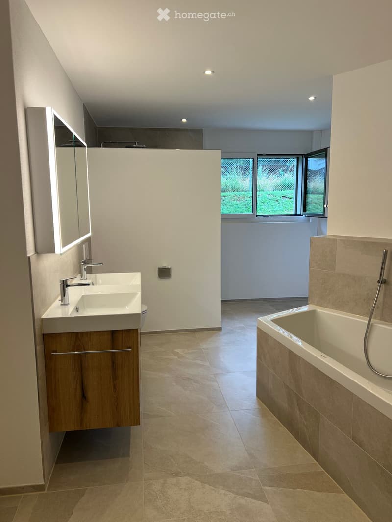 Modern bathroom with a large sink, faucet, bathtub, and a mirrored cabinet with built-in lighting. The space features large windows with a view of greenery outside.