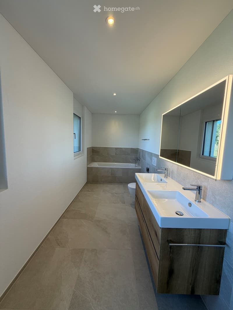 Modern bathroom with double sink, mirror, bathtub, and toilet. Light wood vanity with two sinks and chrome faucets. Neutral tile flooring and walls.