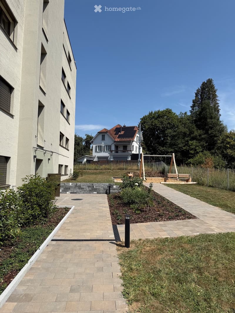 A paved pathway leads to a modern building with a landscaped garden area, featuring shrubs, a bench, and a small playground with swings.