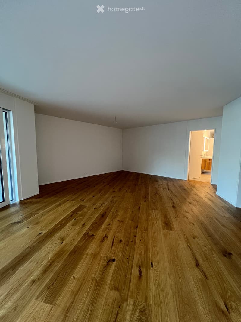 Empty room with light-colored walls and polished wooden flooring, featuring a large window on one side and an open doorway leading to a bathroom.