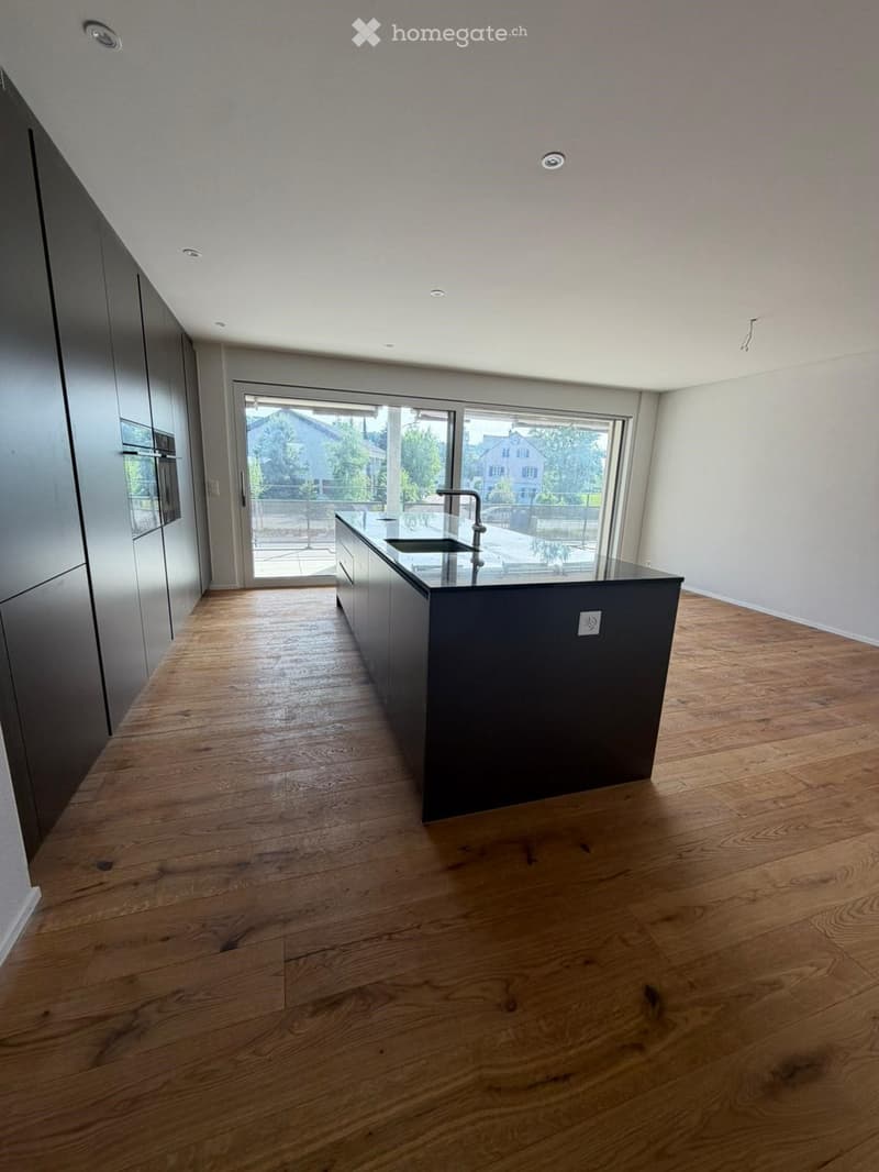 Modern, unfurnished kitchen with wooden flooring, dark grey cabinetry, and a central island with a sink and faucet. Large windows provide natural light and a view of the outdoor area.
