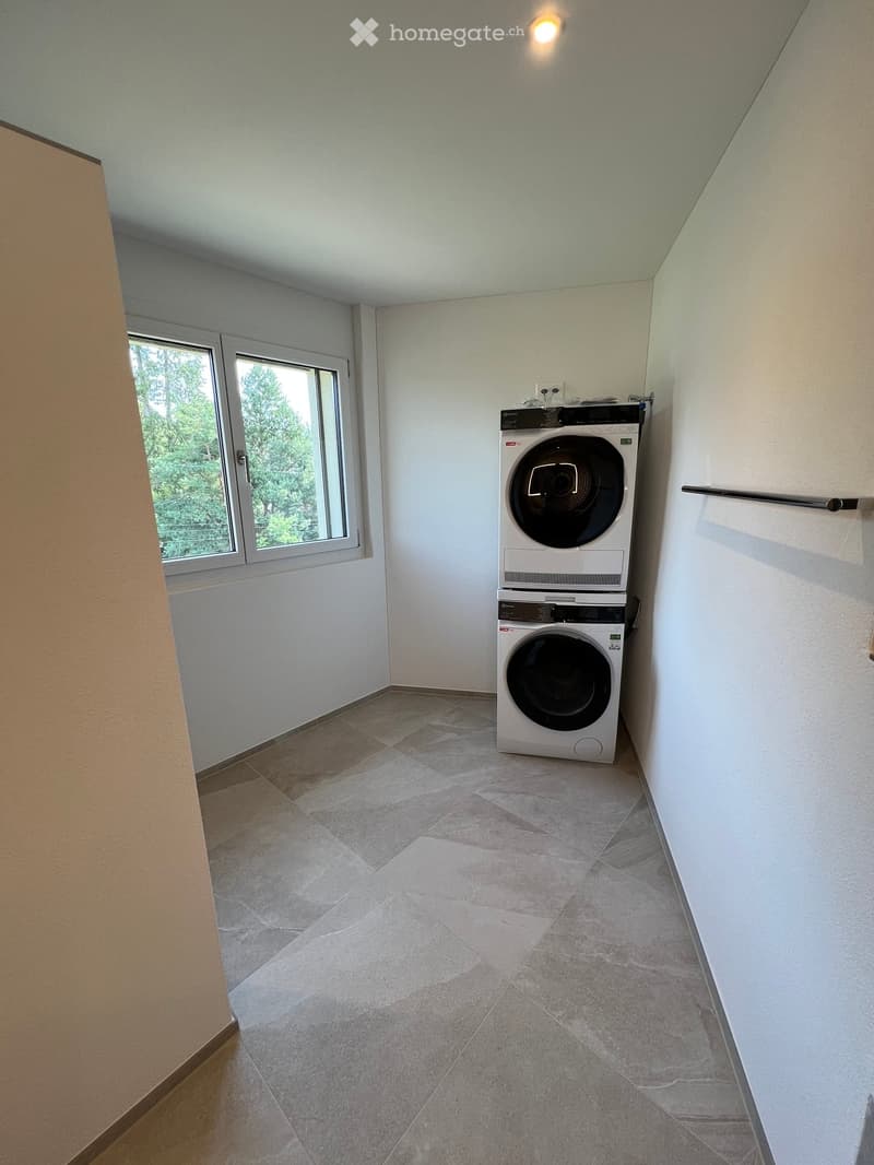 A small, well-lit room with tiled flooring and white walls, featuring a stacked washing machine and dryer unit near a window with a view of greenery.