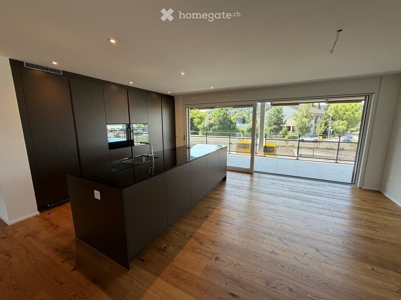 Modern kitchen with dark cabinets, a black countertop, and a wooden floor. Features a large window and sliding glass doors leading to a balcony.