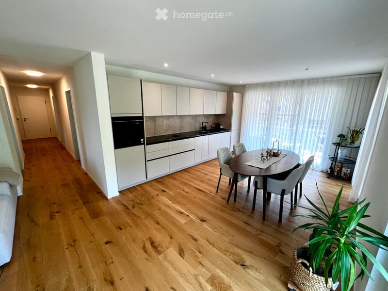 Open-plan kitchen and dining area with light wood flooring, white cabinets, a round dining table with chairs, and a potted plant near the window.