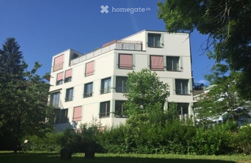 Modern multi-story apartment building with white facade, multiple windows, and balconies. Surrounded by greenery and trees.