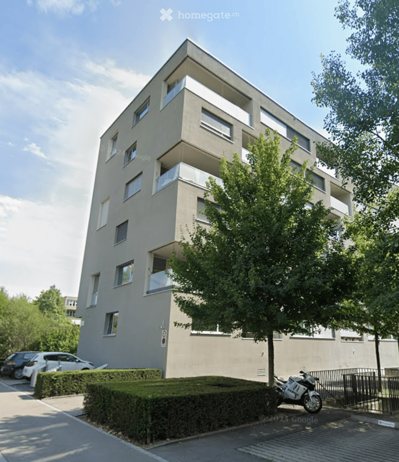 Modern multi-story apartment building with balconies, surrounded by greenery and a parking area.
