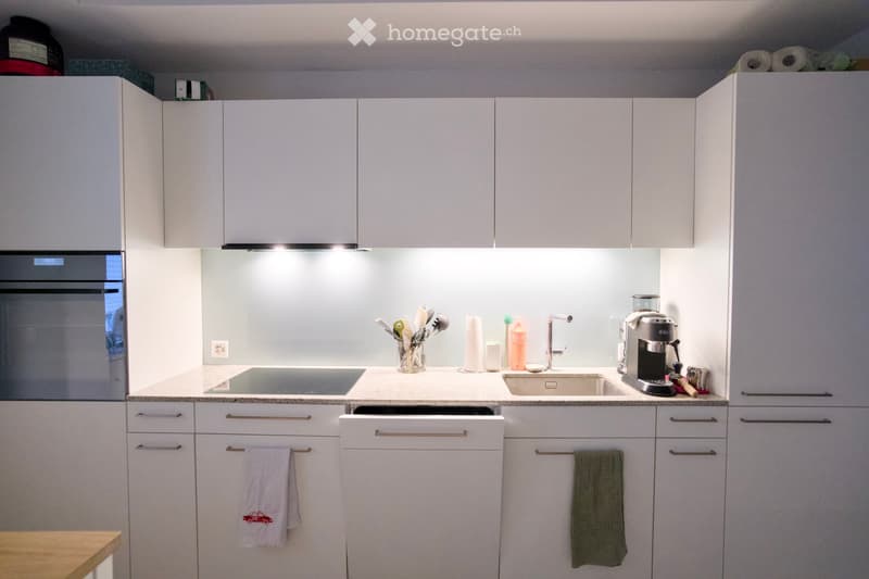 Modern kitchen with white cabinets, stainless steel appliances, and a light-colored countertop. Features include an oven, sink, coffee maker, and a dishwasher.